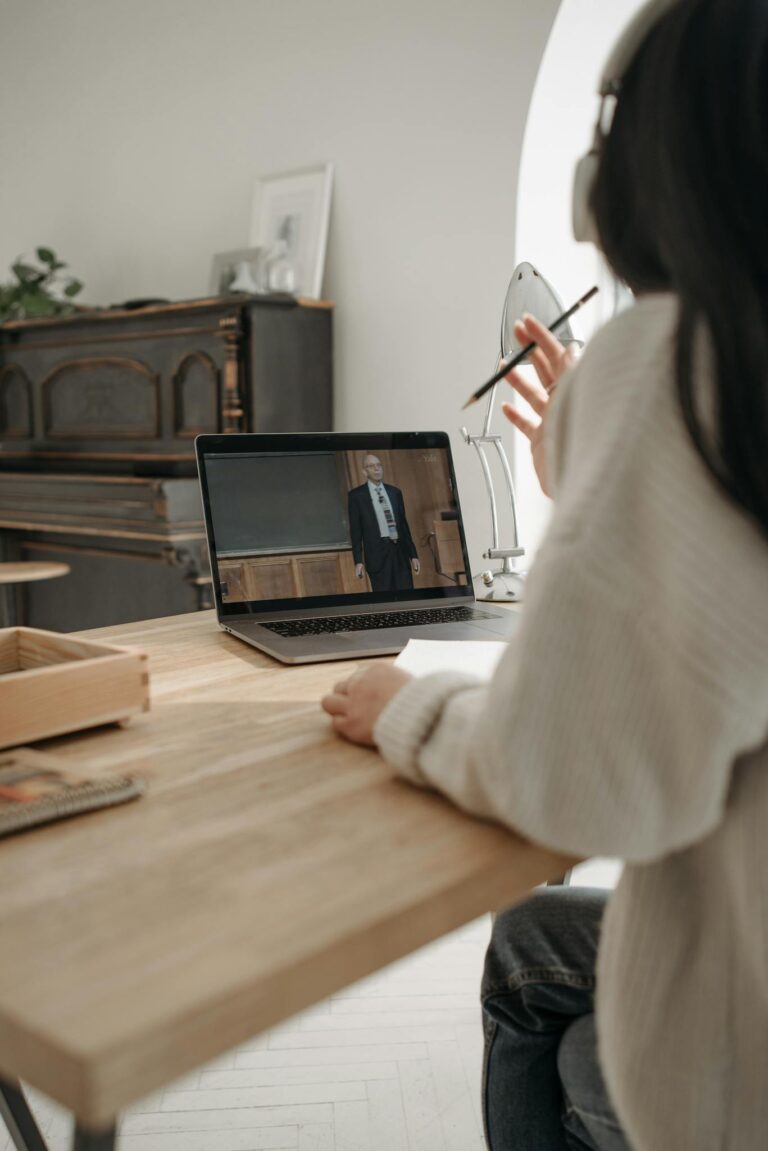 Person attending an online class with a laptop at home, wearing headphones and holding a pen.